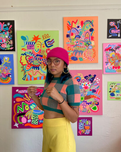 Nikita Ares stands in front of a wall of her artwork, each frame outlined by a different vibrant color - black, blue, green, maroon, orange, pink, etc. Nikita is wearing a pink beanie, yellow pants, a checkered sweater, and red glasses. 