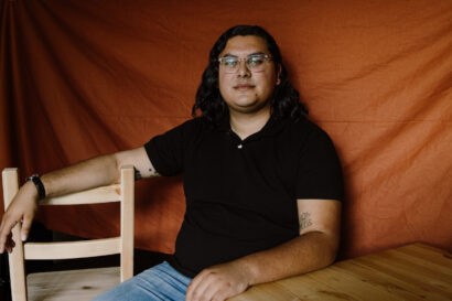 Coby is sitting down wearing a black polo and blue jeans, against a burnt orange background.