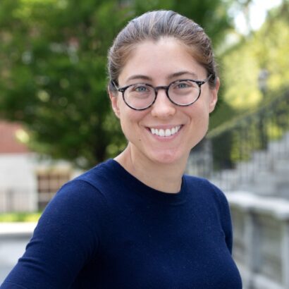 Emily is wearing a Navy Blue top, with glasses on, as she smiles for the photographer. The background is a blurred visual of stone steps, a banister, and a fairly large tree.