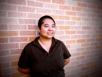 Ching in Chen stands in front of a brick wall, smiling, wearing a black polo.