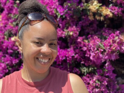 Kimberly is wearing a top that is slightly red and is smiling, head slightly tilted as sun glasses rest atop of Kimberly's head. The background is shrouded with a bushel of vibrant purple flowers.