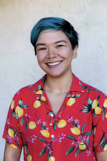 Poet Ally Ang standing in front of white background in a red shirt with blue hair