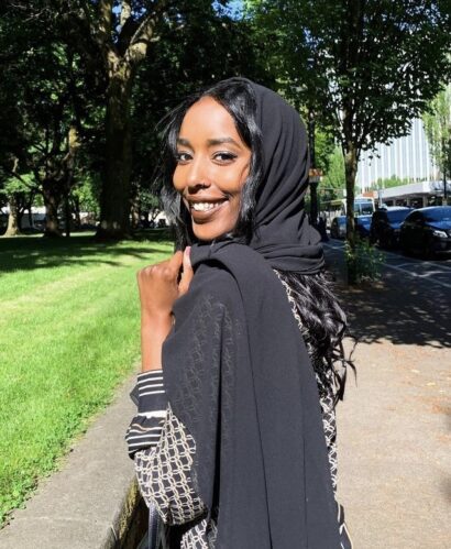 Jamila is standing, smiling at the camera, wearing a black hijab and a black jacket draped over her shoulder on a sunny spring day.