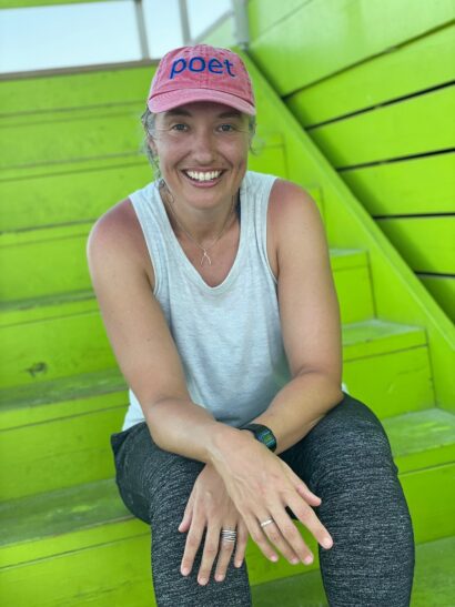 Julie is sitting on a green staircase, smiling, looking directly at the camera. She is wearing a pink hat that says "Poet" and a blue tank top.
