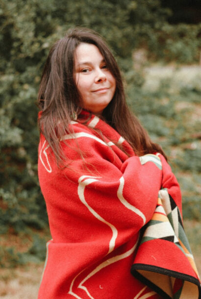 Artist Savannah LeCornu smiling with light brown hair, wearing a blanket around their shoulders