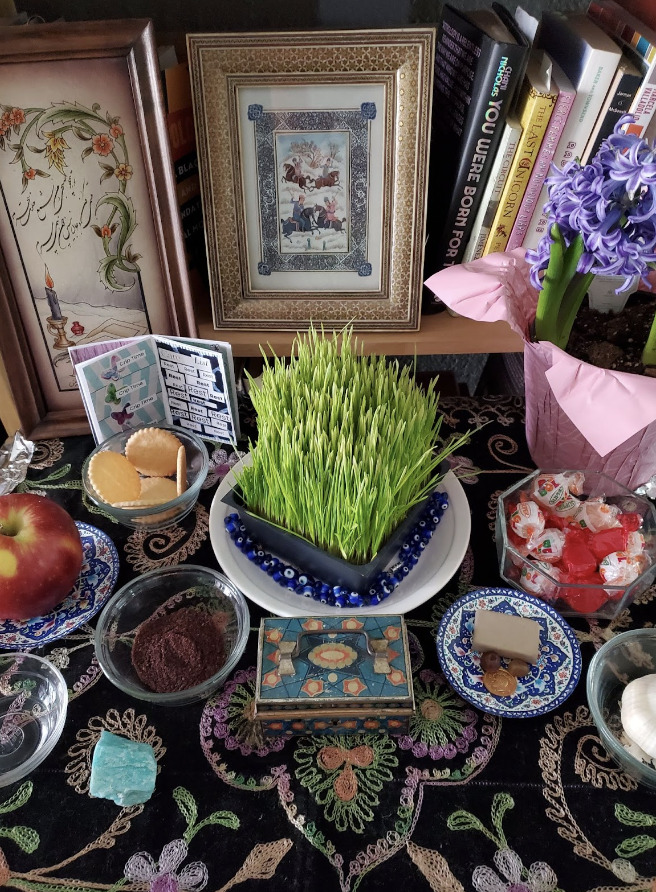 Image Description: A picture of a haft seen, a spring altar for the Iranian New Year. Against a suede tablecloth with embroidered, colorful flowers are little plates and bowls with cookies and candy for sweetness, an apple for beauty, garlic for health, coins for wealth, vinegar for patience, and somac for new beginnings. Green grass is at the center, a symbol of rebirth and growth. In the background of the altar are framed Iranian miniatures. This is my representation of home as love, as ritual.