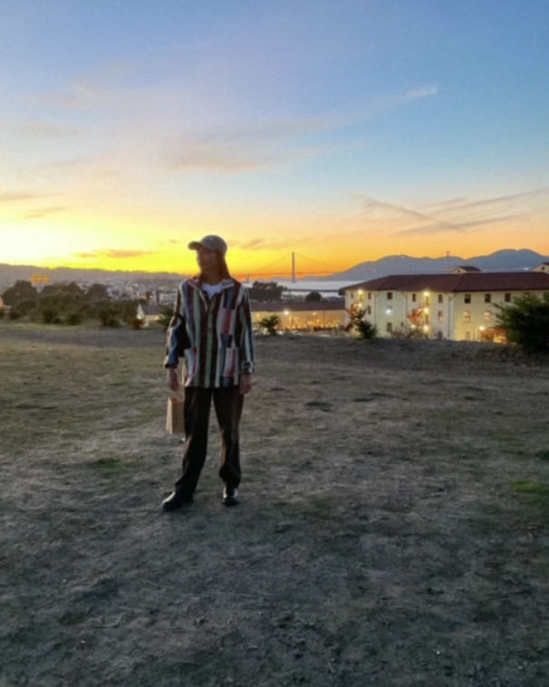 Image Description: Nicole, a brown femme with straightened hair,  stands in a field of short beige grass and dirt, wearing a colorful striped chore jacket, brown pants, black clogs and a grey cap, and holds a small paper shopping bag. The golden gate bridge and bay melt into the sunset in the background; Nicole is not prominent in the picture because she fades into the vibrant orange sunset and dark field. 
