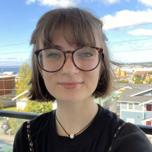 Ally with glasses and short hair smiling wearing a black shirt in front of a blue sky.