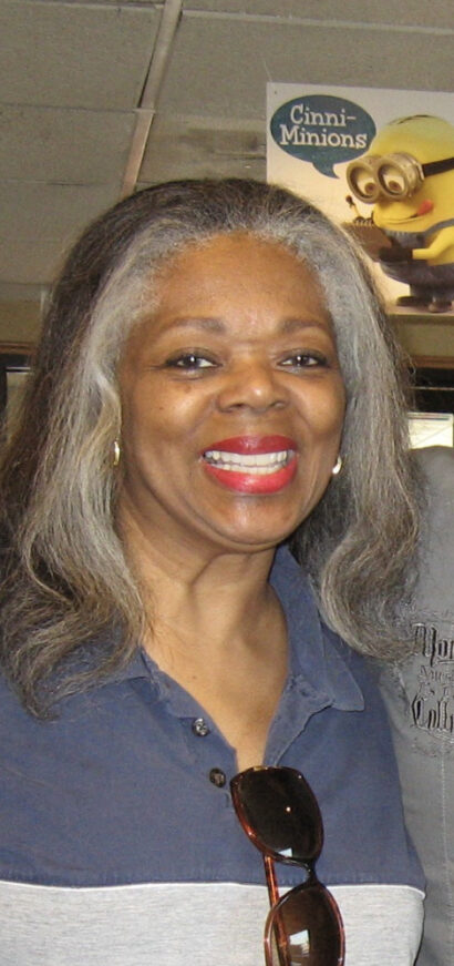 Rose Maria Woodson with grey hair and red lipstick smiling, wearing a blue shirt and sunglasses.