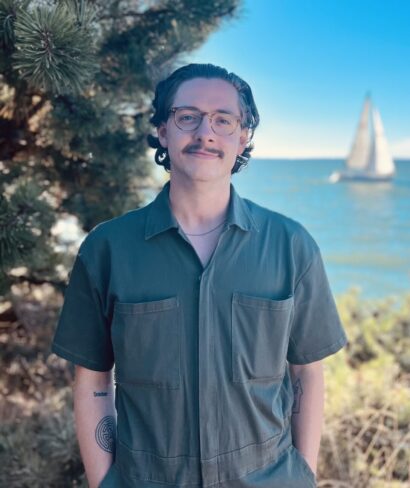 Former ME and writer Sean Dolan smiling in glasses in a green shirt, in front of a water and a boat.