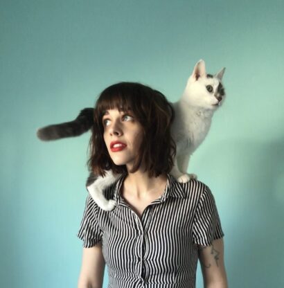 Poet Paige Lewis with short brown hair in a button down shirt and a white cat on their shoulders