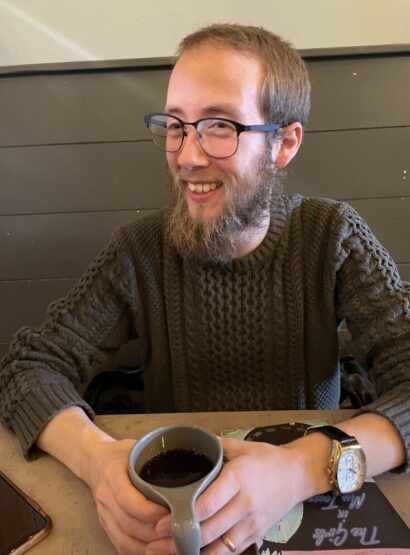 Remi with glasses and a beard, wearing a brown shirt, with a mug of coffee
