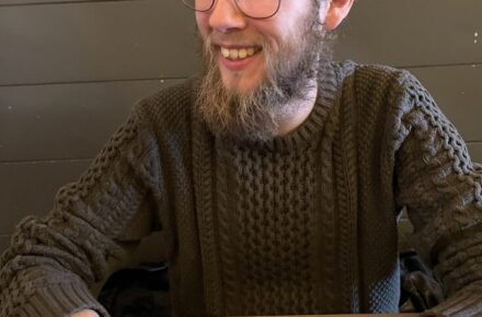 Remi with glasses and a beard, wearing a brown shirt, with a mug of coffee