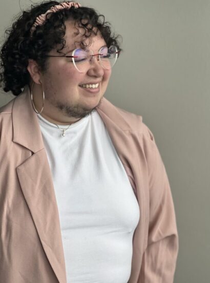 Dallas in round glasses with brown hair smiling in a light pink blazer and white shirt