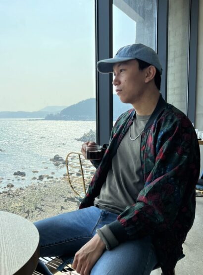 Herie in a baesball cap in front of a beach wearing a multicolored sweater