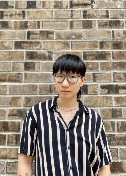 Karen with short black hair and glasses in front of a brick wall, wearing a black and white striped shirt