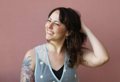 Kira with brown hair and tattoos smiling in front of a mauve background