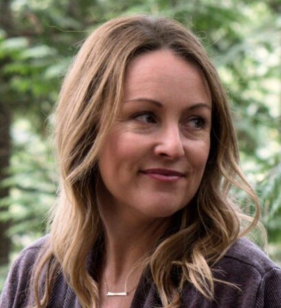 Theresa with blonde hair smiling in front of green foliage