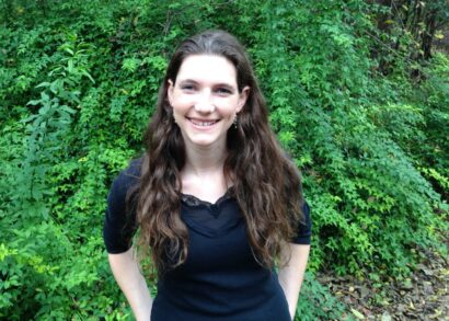 Allison stands in front of a green bush smiling and wearing a black shirt.