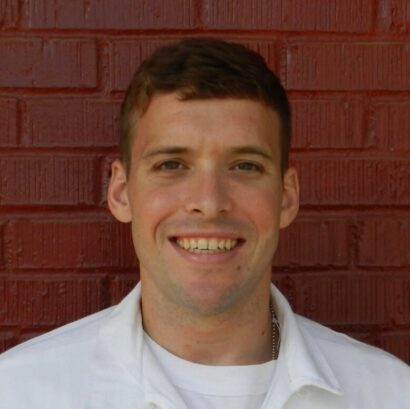 Charles Hill smiles at the camera in a white shirt against a red brick background.