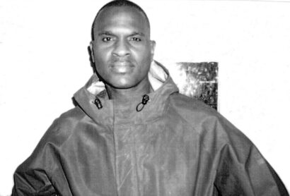 Johnny Lynch looks directly at the camera, wearing a jacket dark jacket against a white backdrop in a black and white photo.