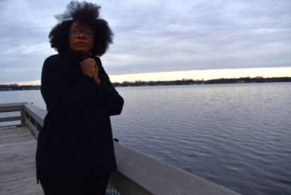 LaToya stands near a body of water clasping her black coat around her against the wind.