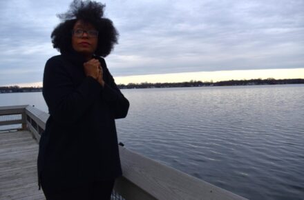 LaToya stands near a body of water clasping her black coat around her against the wind.