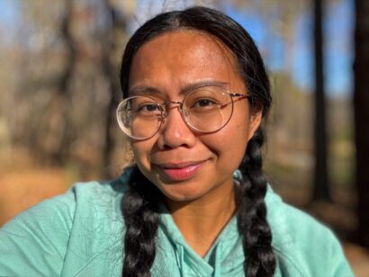 Caroliena looks into the camera. She wears round wire-rimmed glasses and a green shirt. Her black hair falls in pigtails at her shoulders.