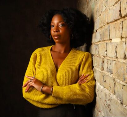 Stacy-Ann in a yellow sweater leaning against a wall in a dim studio