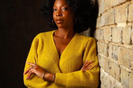 Stacy-Ann in a yellow sweater leaning against a wall in a dim studio