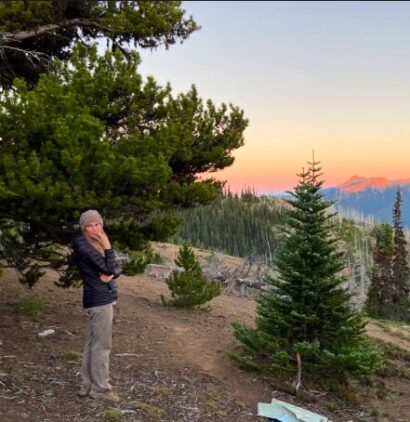 Annie standing in a puffer coat overlooking a view of the sun setting onto a mountain range