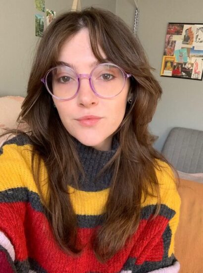 Flannery Maeve Rollins smiles slightly at the camera against the backdrop of a mostly gray room. She wears large round glasses, a striped yellow and red sweater, and her hair is long and brown.