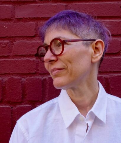 Photo of Emma Goldman-Sherman against a red brick backdrop. They are wearing round red glasses and a white button-up shirt, and have short purple hair. They are looking to the left and are smiling slightly.