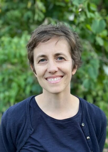 Sonja Sharp looks directly at the camera and smiles against a background of green leaves. She is wearing a dark blue top, a dark blue cardigan that is unbuttoned, and she has short brown slightly messy hair.