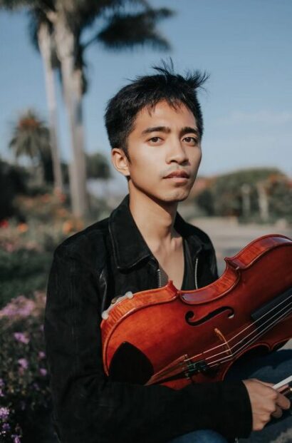 Jay Julio, a person with short black hair, gazes into the camera against a backdrop of a sunny tropical location. They are wearing a silky black button-up shirt and are holding a reddish-brown viola.