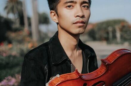 Jay Julio, a person with short black hair, gazes into the camera against a backdrop of a sunny tropical location. They are wearing a silky black button-up shirt and are holding a reddish-brown viola.