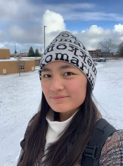 Tuuli Qin-Terrell smiles at the camera against a wintry backdrop. She is wearing a white beanie hat with black letters on it, a white turtleneck, and a red and gray patterned jacket.