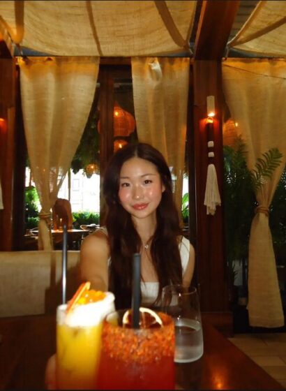 Photo of Zixuan Angel Xin wearing a white tank top and red lipstick smiling at a dimly-lit dinner table