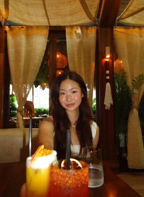 Photo of Zixuan Angel Xin wearing a white tank top and red lipstick smiling at a dimly-lit dinner table