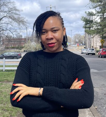 The author Obiageli, a woman wearing red lipstick and nail polish, folds her arms and looks into the camera