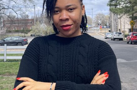 The author Obiageli, a woman wearing red lipstick and nail polish, folds her arms and looks into the camera