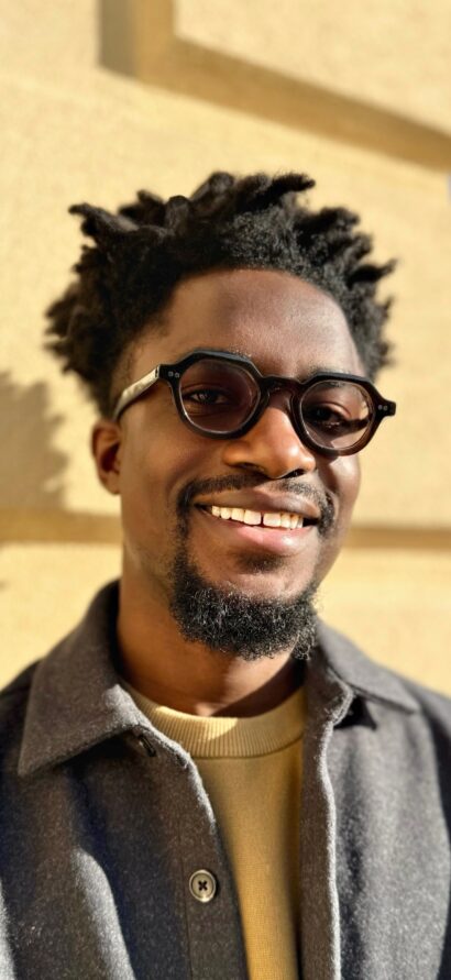 Author Dolapo Demuren smiles at the camera in sunlight. He is wearing round glasses and a grey jacket.