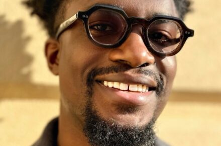 Author Dolapo Demuren smiles at the camera in sunlight. He is wearing round glasses and a grey jacket.