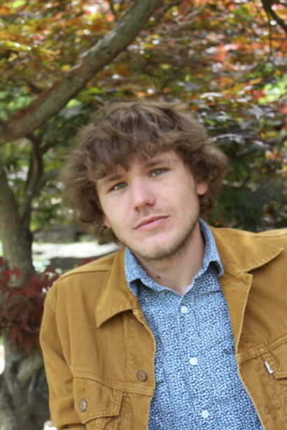 Author Brock Storey, a man with short brown hair, looks at the camera with a tree in the sunny background
