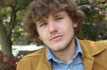 Author Brock Storey, a man with short brown hair, looks at the camera with a tree in the sunny background