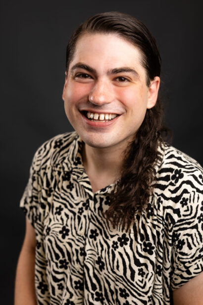 Author Joshua Zeitler, a person with long brown hair wearing a patterned black and white buttoned shirt, smiles widely at the camera