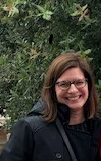 Author Kelleen Zubick, a woman with brown hair and glasses, smiles widely against a backdrop of a tree