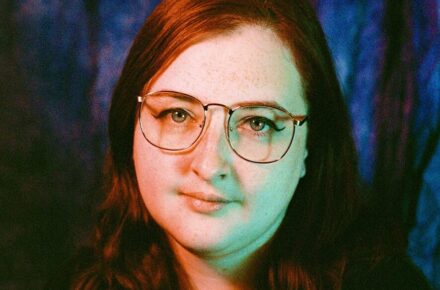 Nat with red hair and glasses in front of a blue background