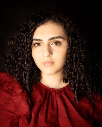 Author Raya Tuffaha, a woman with dark curly hair wearing a red blouse, looks pensively into the camera