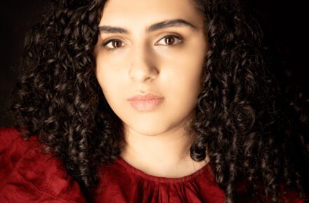 Author Raya Tuffaha, a woman with dark curly hair wearing a red blouse, looks pensively into the camera